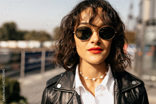 Bright curly woman with red lips in sunglasses in black jacket and white blouse looking at camera . Fashionable girl in good mood smiling outdoor