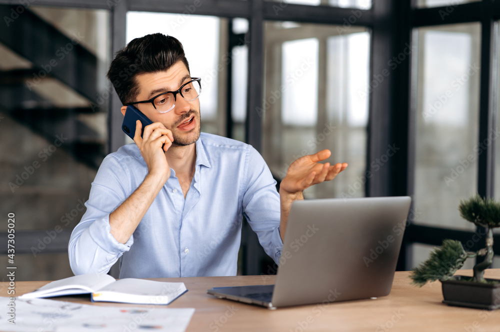 Confident caucasian freelancer talking on smartphone while sitting at desk in modern office at а laptop, successful man in strict shirt, have phone conversation, gesturing with his hand Confident caucasian freelancer talking on smartphone while sitting at desk in modern office at а laptop, successful man in strict shirt, have phone conversation, gesturing with his hand