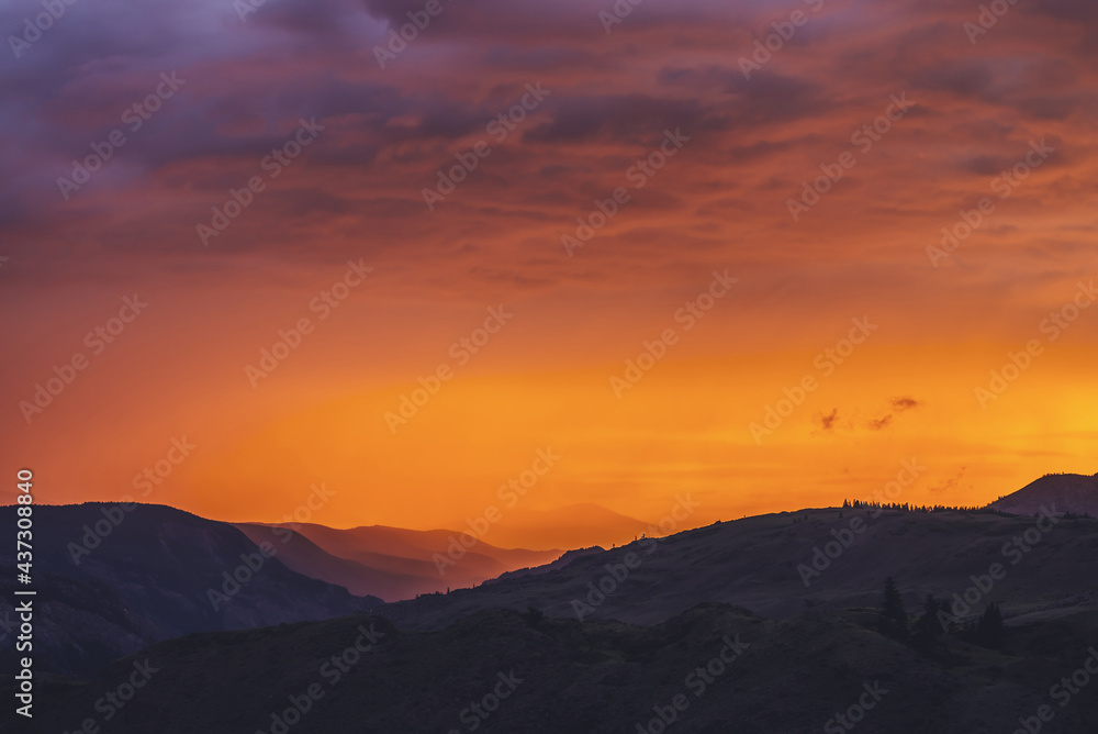 Atmospheric landscape with silhouettes of mountains with trees on background of vivid orange pink violet dawn sky. Colorful nature scenery with sunset or sunrise of illuminating color. Sundown paysage