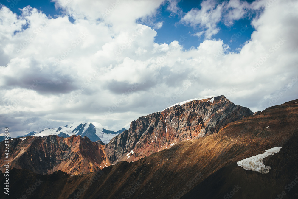 Fototapeta premium Scenic highlands landscape with great snowy mountain peak behind colorful brown red orange mountain wall in sunlight. Sunny mountains scenery with high vivid brown red orange mountain and big snow top