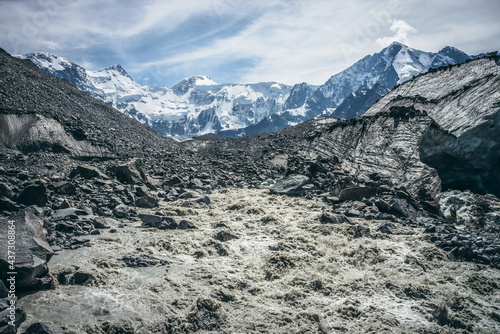 Scenic landscape with powerful mountain river beginning from glacier among large moraines on background of great snowy mountains. Beautiful scenery with glacier at source of turbulent mountain river.