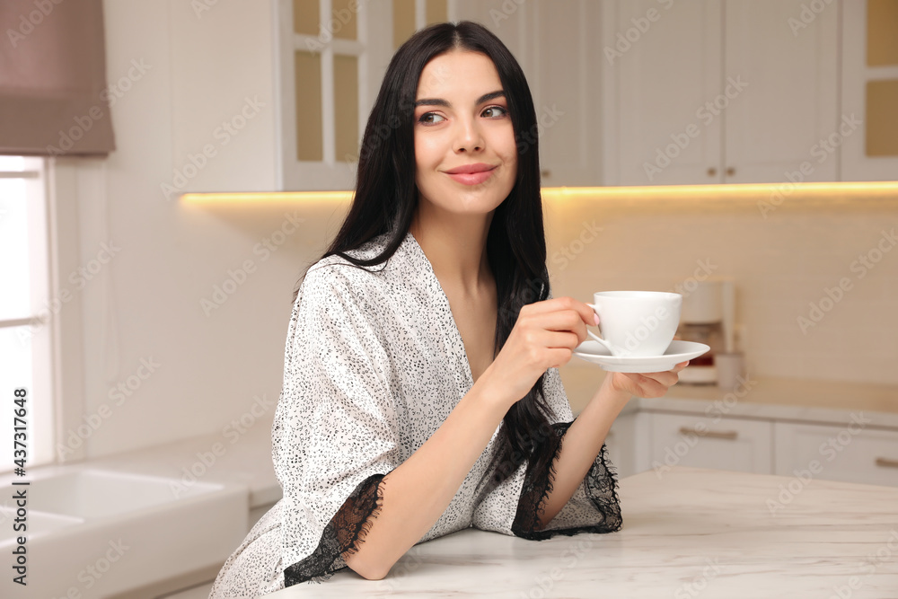 Pretty young woman wearing beautiful silk robe with cup of coffee in kitchen
