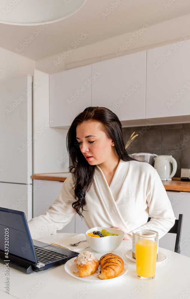 Young woman in comfortable white robe works on laptop while sitting at table the kitchen. Work at home for breakfast. Young woman in comfortable white robe works on laptop while sitting at table the kitchen. Work at home for breakfast.
