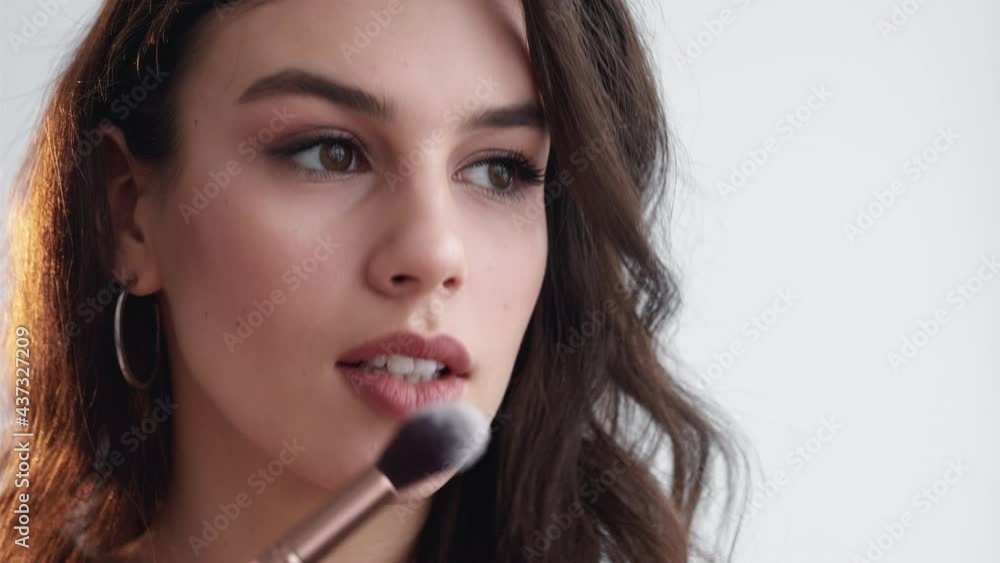 Facial makeup. Visage tutorial. Beauty look. Brunette woman applying blush with powder brush on flawless radiant face skin cheek isolated on white background. Facial makeup. Visage tutorial. Beauty look. Brunette woman applying blush with powder brush on flawless radiant face skin cheek isolated on white background.