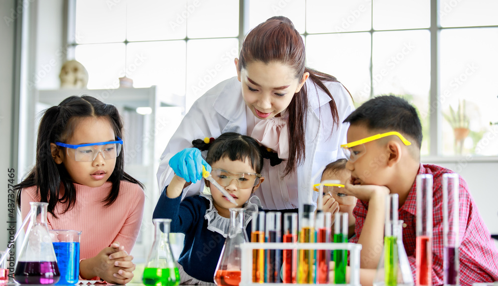 Asian little boy drop reagent sample by glass dropper into colorful solution flask while young girls wears safety glasses and teacher in white lab coat look at him do experiment in school laboratory. Asian little boy drop reagent sample by glass dropper into colorful solution flask while young girls wears safety glasses and teacher in white lab coat look at him do experiment in school laboratory.