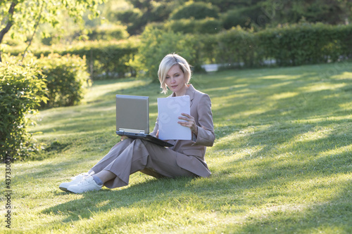 Woman working in the park with a laptop in the summer in the sunbeams. she has a notebook in her hands.