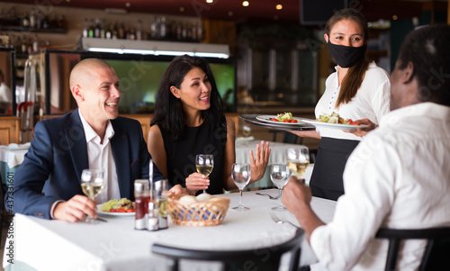 Young waitress in protective face mask serving meals to friendly company in restaurant. Precautions in catering establishments during pandemic coronavirus