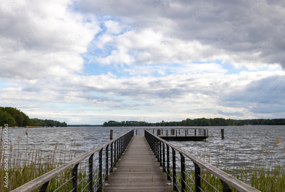 a long wooden jetty by a lake. a bright blue sky with many clouds in the sky.