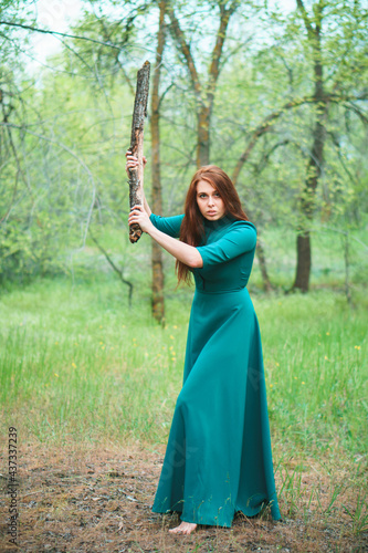 Young woman in elegant dress with cudgel in hands. Lady in spring forest. Danger and self-defence concept.