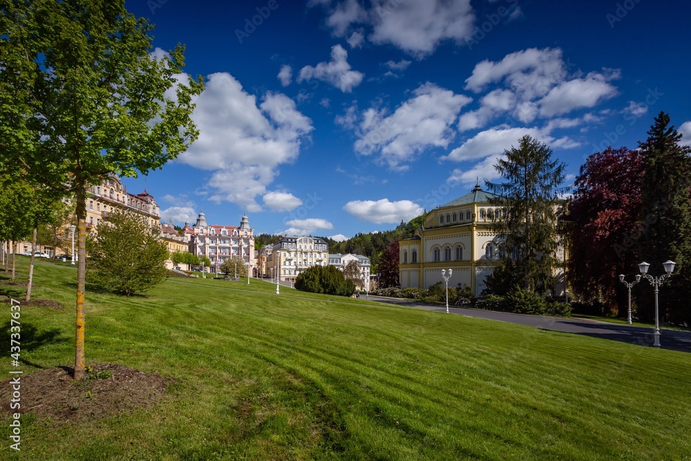 Marianske Lazne, Czech Republic - May 30 2021: View of the Goethe square with hotel buildings and a church around. Green lawn and trees in the foreground. Sunny day with blue sky and clouds. Spa city.