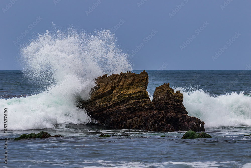 Ocean wave breaking on rock just offshore in Laguna Beach, California. White spray in the air;  blue sky in background. 
