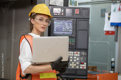 Factory worker is programming a CNC milling machine with a tablet computer. engineering and worker woman in safety hard hat and reflective cloth using lathe machine inside the factory.