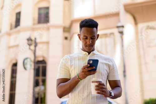 Portrait of young African businessman wearing casual clothes while texting with mobile phone and holding takeaway coffee cup