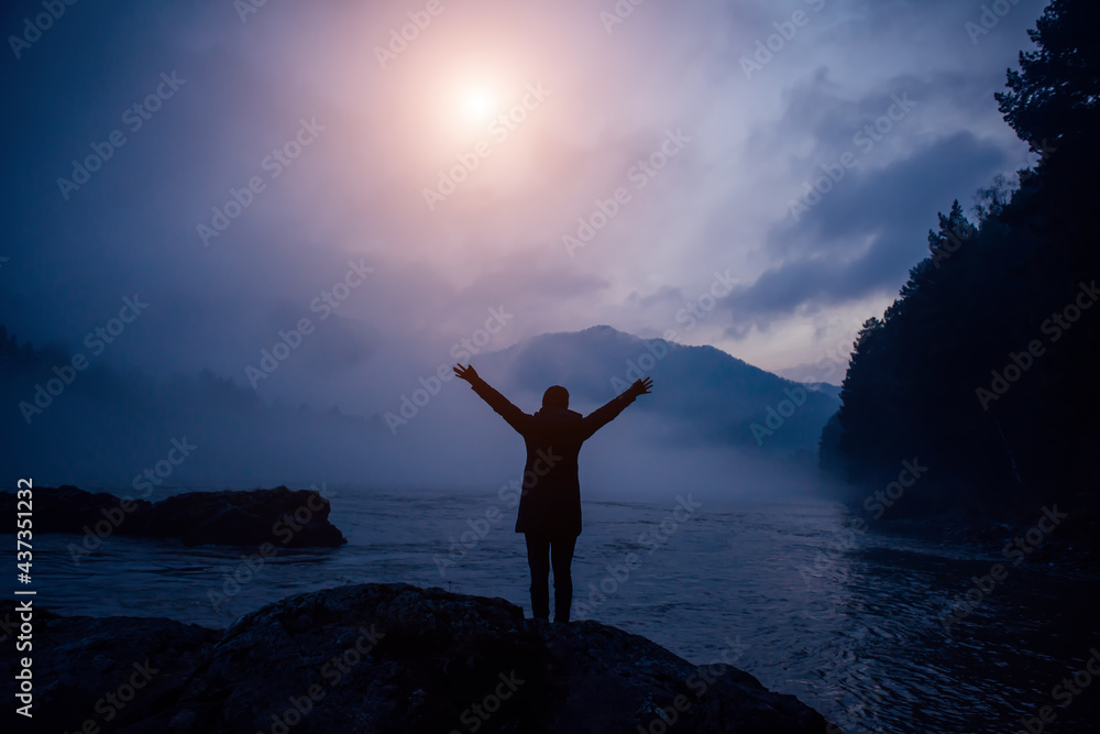 Blue evening mist over mountain river on the background of wooded hills and overcast sky. Man stands in the fog, hands up, looking at the setting sun. Woman's appeal to the power of nature.