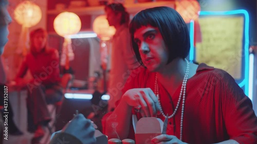 Over the shoulder shot of transgender man in wig and female outfit speaking with friend, eating noodles and drinking from paper cup while sitting in Chinese restaurant photo