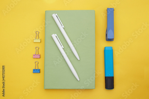 Top view of green book , white pen and school suppliers on yellow background 