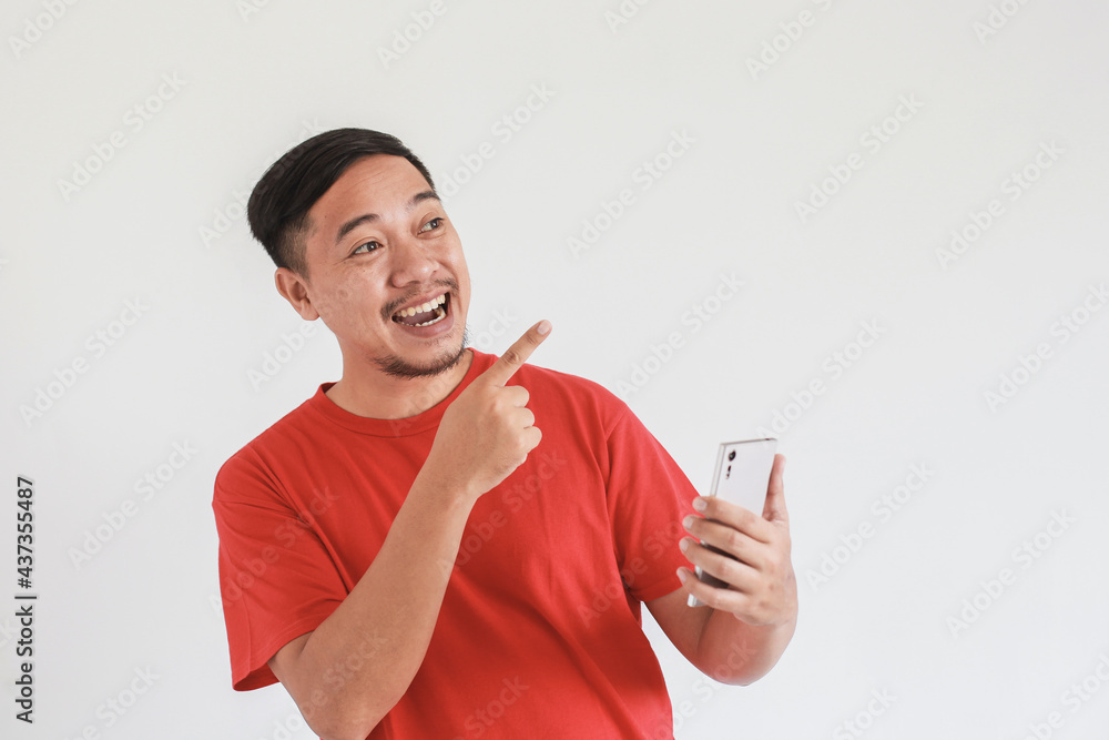 Smiling face expression of happy Asian man wearing red t-shirt with index finger pointing to empty space while using smart phone Smiling face expression of happy Asian man wearing red t-shirt with index finger pointing to empty space while using smart phone