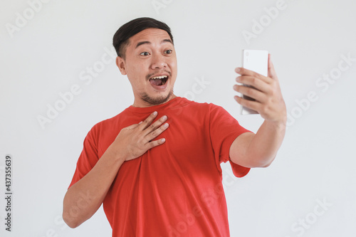 Surprised and shocked Asian man in red t-shirt using smartphone on white background