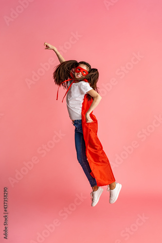 Fototapeta Naklejka Na Ścianę i Meble -  beautiful smiling girl in superhero costume, in red cape and mask shows how strong she is, isolated on a pink background in full growth. Cute baby playing superhero game. concept of Girl Power.