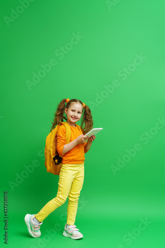 E-learning. cute schoolgirl with two ponytails with backpack and tablet computer in her hands stands on green background, Child smiles and looks into the frame. Recommending a Remote online school.