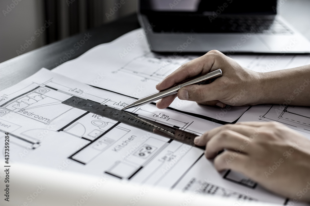 A close-up, an architect, a house designer holding a pen, pointing to a house plan to examine the design plan before discussing the details with the client. Interior design and decoration ideas. A close-up, an architect, a house designer holding a pen, pointing to a house plan to examine the design plan before discussing the details with the client. Interior design and decoration ideas.