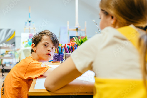 Rear view of nursery teacher sitting with boy child down syndrome looking at camera drawing in the classroom.