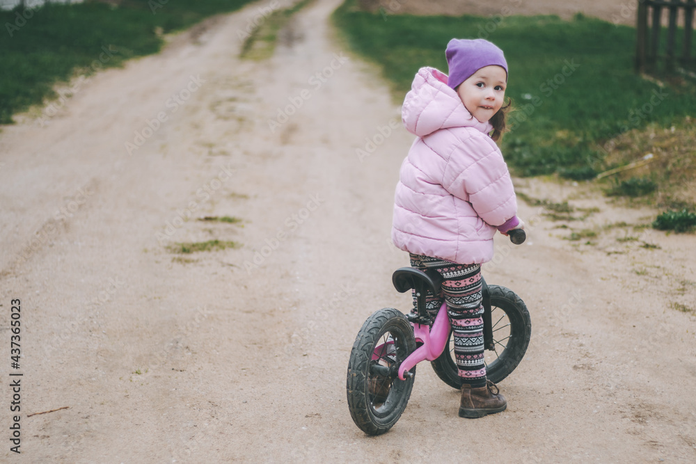 Child in pink jacket and lilac hat rides balance bike on dirt road, in countryside Child in pink jacket and lilac hat rides balance bike on dirt road, in countryside