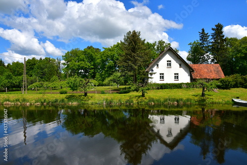 A view of an abandoned house or shack located next to the coast of a river or lake with the bank of the reservoir covered with trees, shrubs, flowers, grass, and reeds seen on a sunny day in Poland