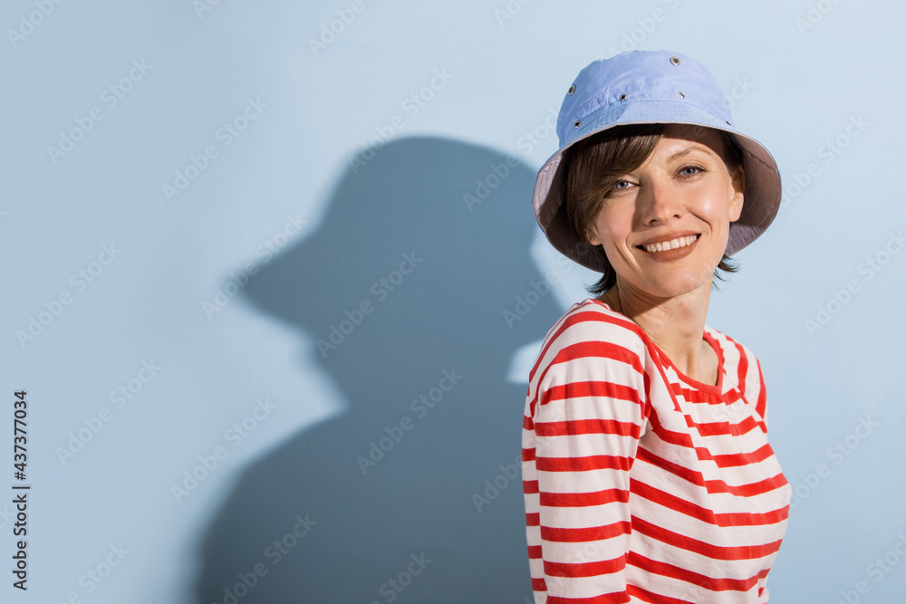 Happy cheerful young woman with short hair, natural makeup over light blue background. Indoor portrait of beautiful brunette young woman wearing blue hat and bright striped clothing Happy cheerful young woman with short hair, natural makeup over light blue background. Indoor portrait of beautiful brunette young woman wearing blue hat and bright striped clothing
