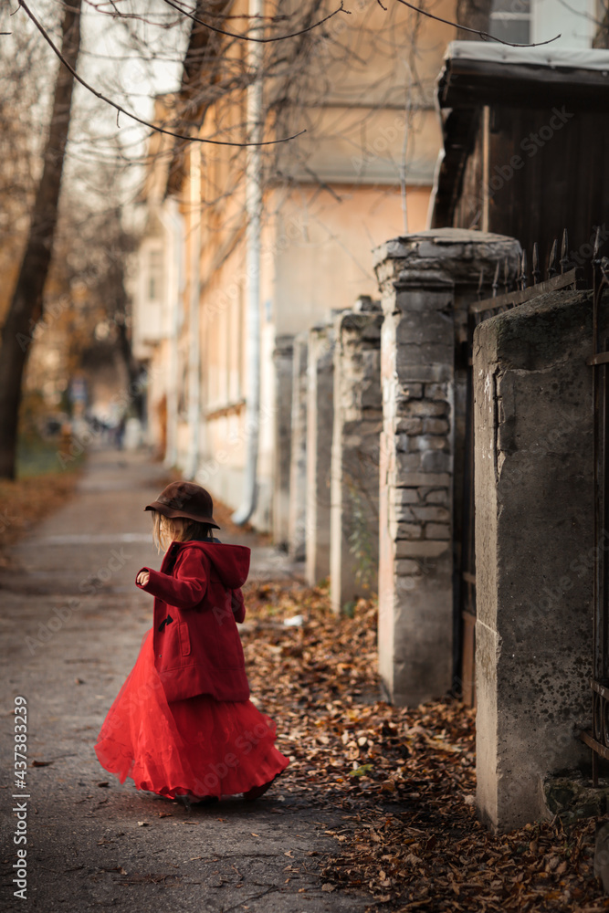Cute funny caucasian child girl in a red coat walks on the autumn street, stylish child, kid girl in a festive dress for a walk