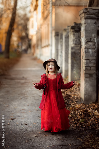 Cute funny caucasian child girl in a red coat walks on the autumn street, stylish child, kid girl in a festive dress for a walk