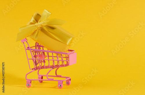 yellow gift box with a silk bow in a miniature metal trolley on a yellow background.