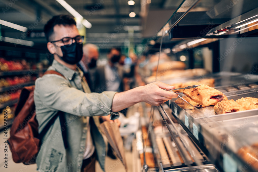 Young man buying freshly baked pastry in big modern bakery. He is wearing face protective mask due to Coronavirus epidemic. Young man buying freshly baked pastry in big modern bakery. He is wearing face protective mask due to Coronavirus epidemic.