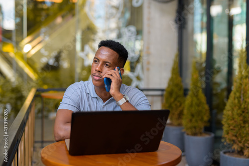 African businessman sitting at coffee shop while using laptop computer and talking on mobile phone