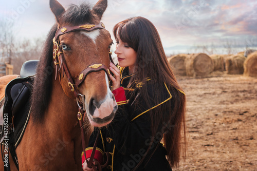 A beautiful woman with long and black hair in a historical hussar costume stands in a field with a horse.