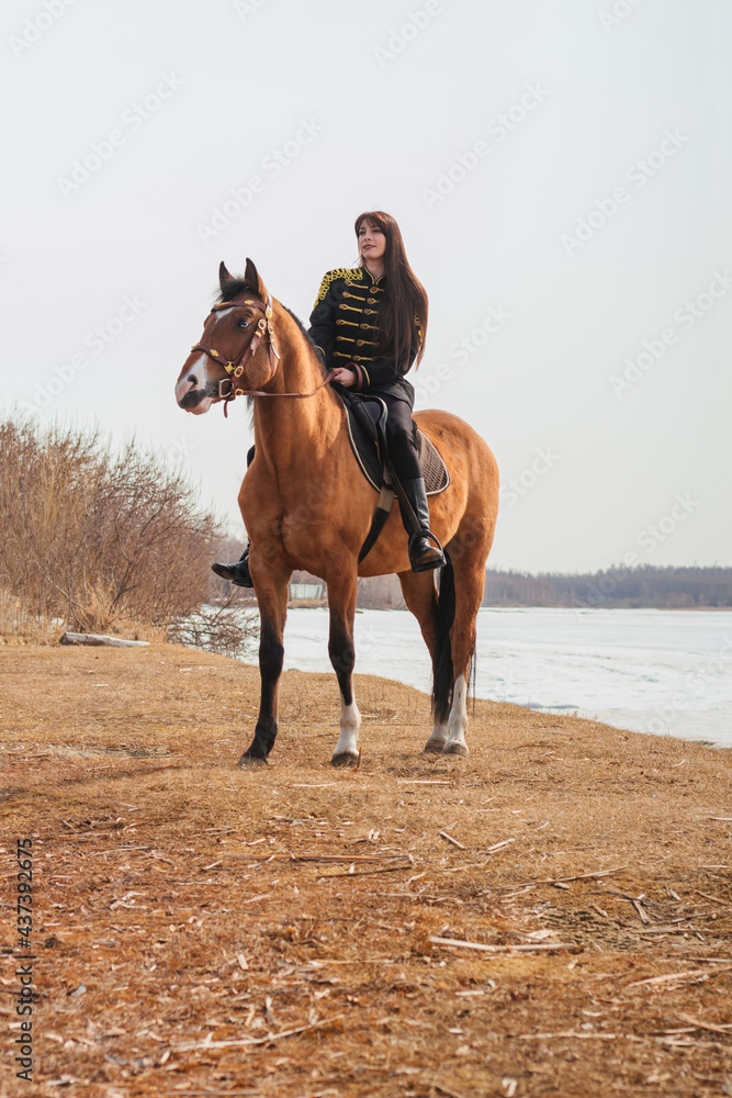 A beautiful woman with long and black hair in a historical hussar costume stands near a river with a horse. A beautiful woman with long and black hair in a historical hussar costume stands near a river with a horse.