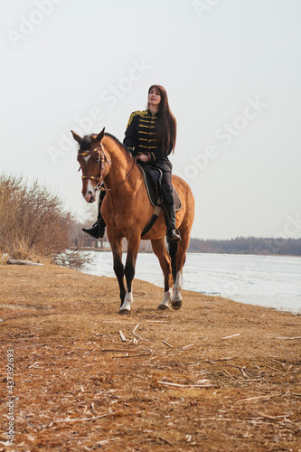 A beautiful woman with long and black hair in a historical hussar costume stands near a river with a horse. © Вероника Преображенс