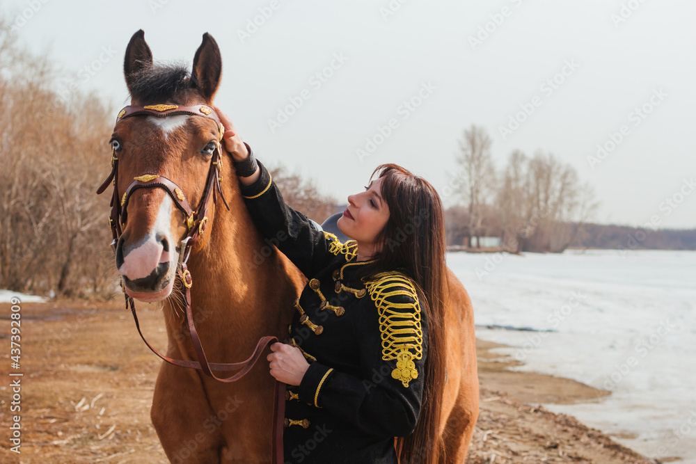 Obraz premium A beautiful woman with long and black hair in a historical hussar costume stands near a river with a horse.