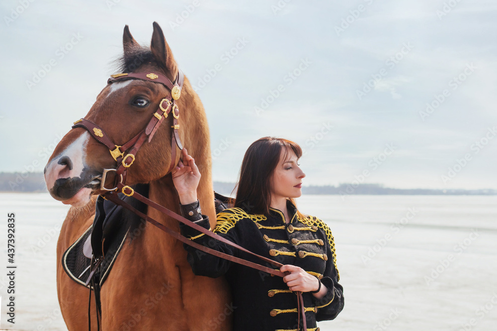 Obraz premium A beautiful woman with long and black hair in a historical hussar costume stands near a river with a horse.