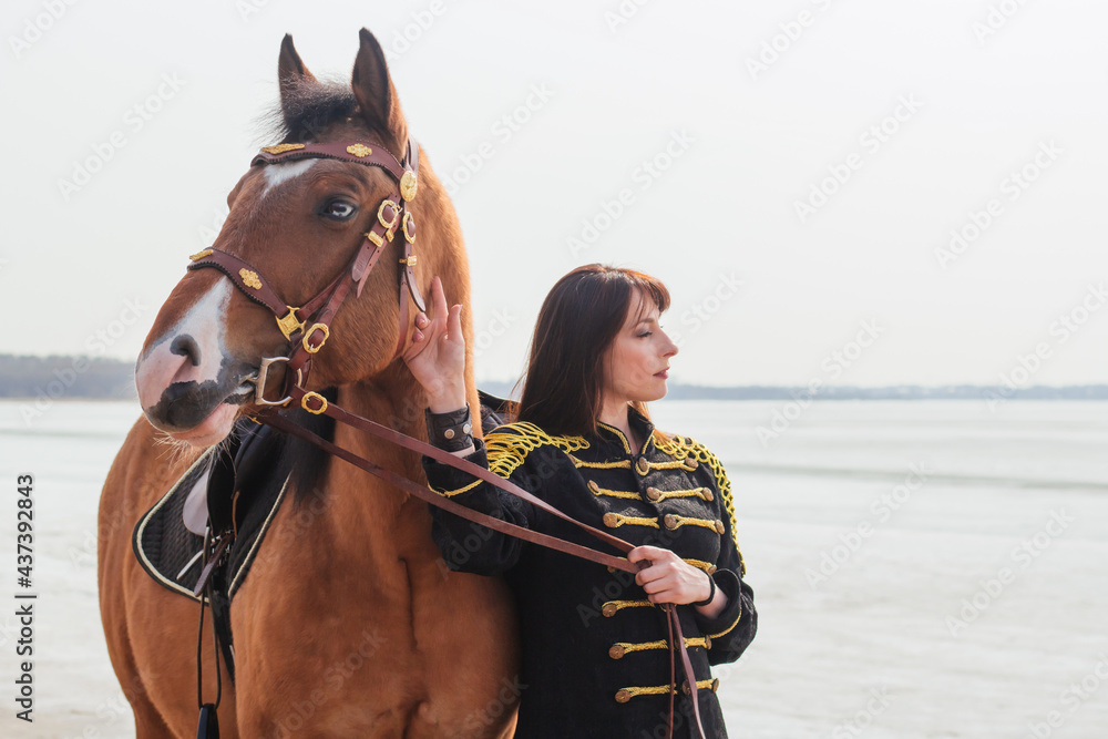 Obraz premium A beautiful woman with long and black hair in a historical hussar costume stands near a river with a horse.