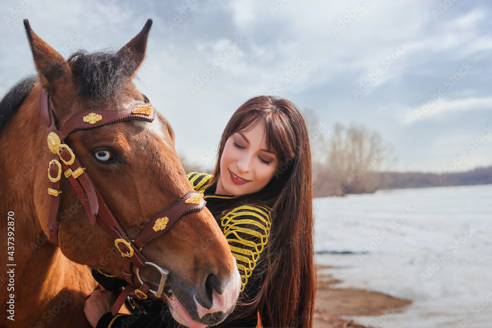 Fototapeta premium A beautiful woman with long and black hair in a historical hussar costume stands near a river with a horse.