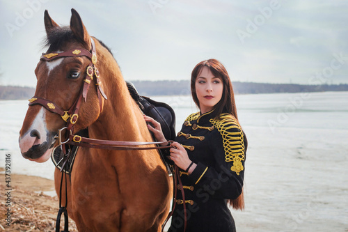 A beautiful woman with long and black hair in a historical hussar costume stands near a river with a horse.