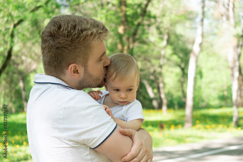 Young caucasian father comforts holds sad baby, kissing among green trees in park in summer. Dad and kid relationship, concept Young caucasian father comforts holds sad baby, kissing among green trees in park in summer. Dad and kid relationship, concept