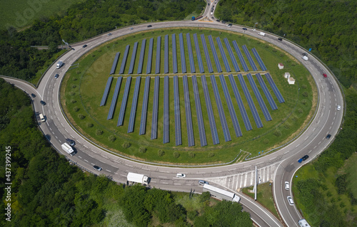 Large roundabout in the forest aerial view. Solar panels in the center of a circular automobile ring. Car traffic on the motorway top view. Italy roads aerial view. Solar panels by the road.