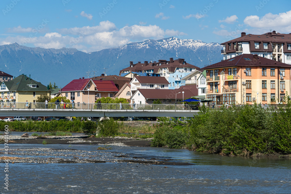 Residential buildings in federal territory of Sirius against backdrop of snow-capped peaks. Caucasus mountains. Imeretinskaya lowland. In foreground is Mzymta.Adler. Sochi, Russia - May 18, 2021
