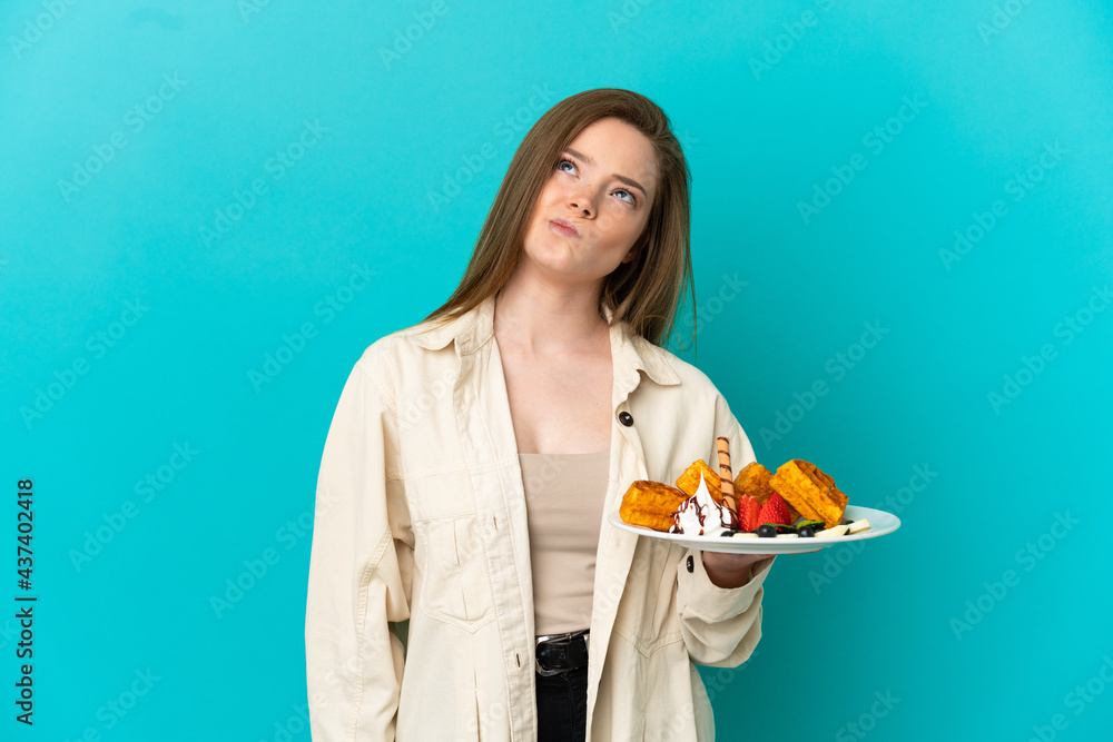 Teenager girl holding waffles over isolated blue background and looking up Teenager girl holding waffles over isolated blue background and looking up