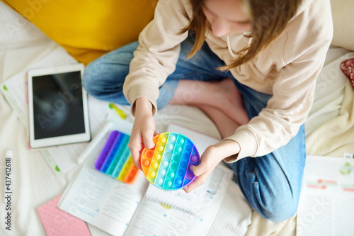 Teenage girl playing with rainbow pop-it fidget toy while studying at home. Teen kid with trendy stress and anxiety relief fidgeting game. photo