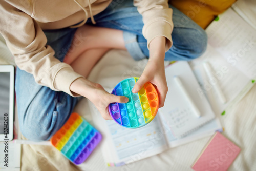 Teenage girl playing with rainbow pop-it fidget toy while studying at home. Teen kid with trendy stress and anxiety relief fidgeting game. photo