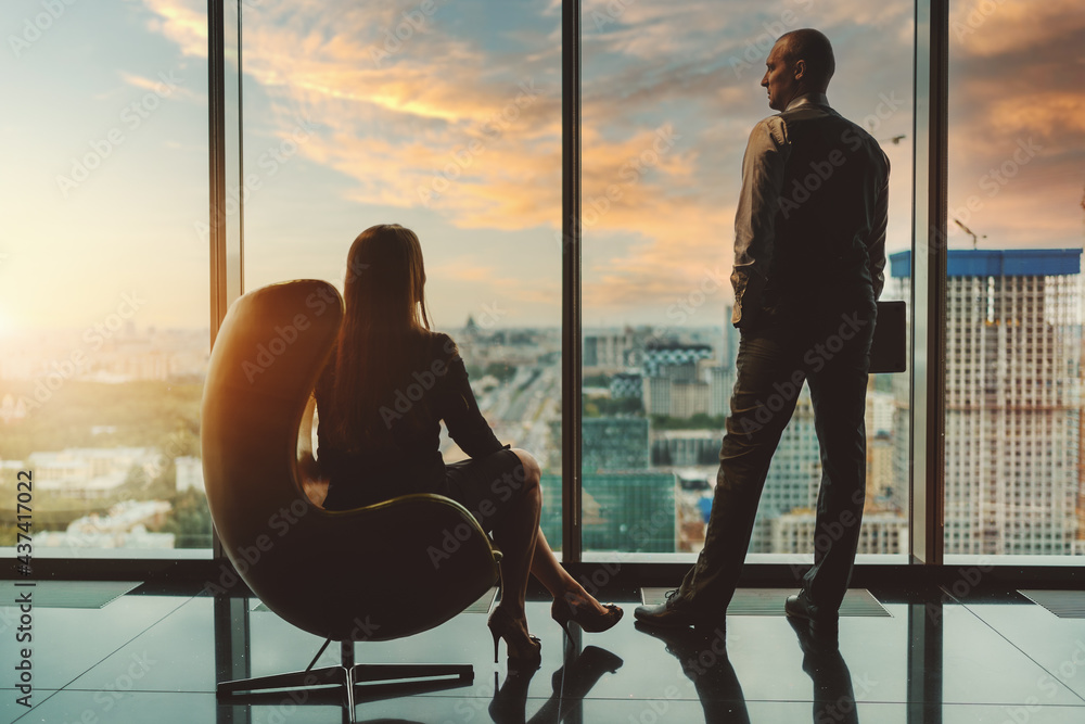 Silhouettes of two entrepreneurs on the top floor of a business high-rise, enjoying the sunset after a hard workday: a businessman standing next to the window, a businesswoman sitting in an armchair