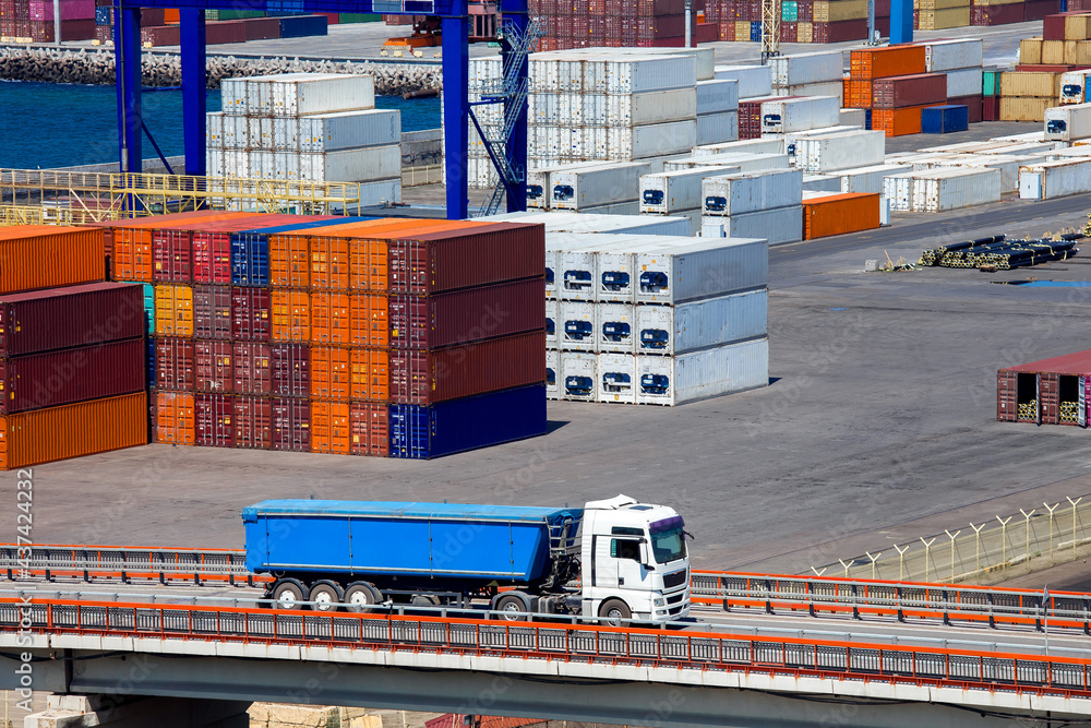 Φωτογραφία truck with a trailer rides over a bridge in the background a warehouse of a merchant port with stacked containers of export goods brought by a ship along the sea route
